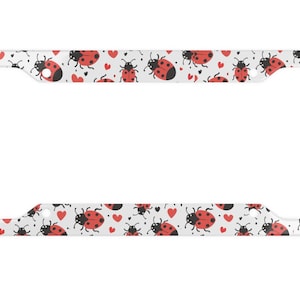 May include: White metal license plate frame with a pattern of red ladybugs and red hearts on a white background.