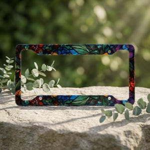May include: A rectangular license plate frame featuring a vibrant, multi-colored stained glass pattern. The design consists of intricate floral and leaf motifs in shades of deep blue, green, red, and orange, with four pre-drilled mounting holes.