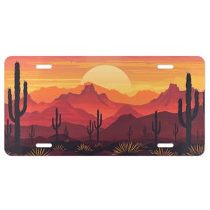 May include: License plate featuring a desert landscape at sunset. The design includes silhouetted cacti, mountains, and a large sun in shades of orange, red, and yellow. The plate has four mounting holes.