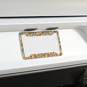 May include: License plate frame with a teal background and a pattern of oranges and white flowers. The frame is rectangular with a white center for the license plate.