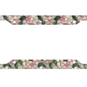 May include: A license plate frame with a floral pattern. The frame has a design of pink and white flowers with green leaves, set against a light background. The design is similar to stained glass, with gold-coloured outlines.