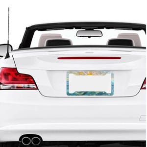 May include: A rectangular license plate frame featuring a stylized artistic print of a sun rising over blue ocean waves. The frame has four mounting holes and is attached to the rear trunk of a white convertible car with red taillights.