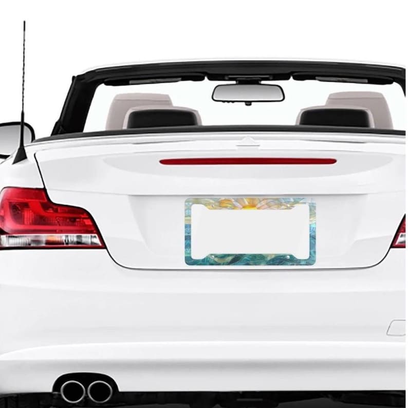 May include: A rectangular license plate frame featuring a stylized artistic print of a sun rising over blue ocean waves. The frame has four mounting holes and is attached to the rear trunk of a white convertible car with red taillights.