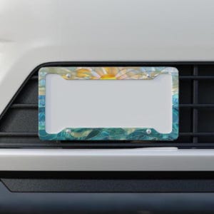 May include: A rectangular license plate frame featuring a stained glass style pattern. The design displays a yellow sun rising over blue and teal waves, with four silver mounting screws visible at the corners of the frame.