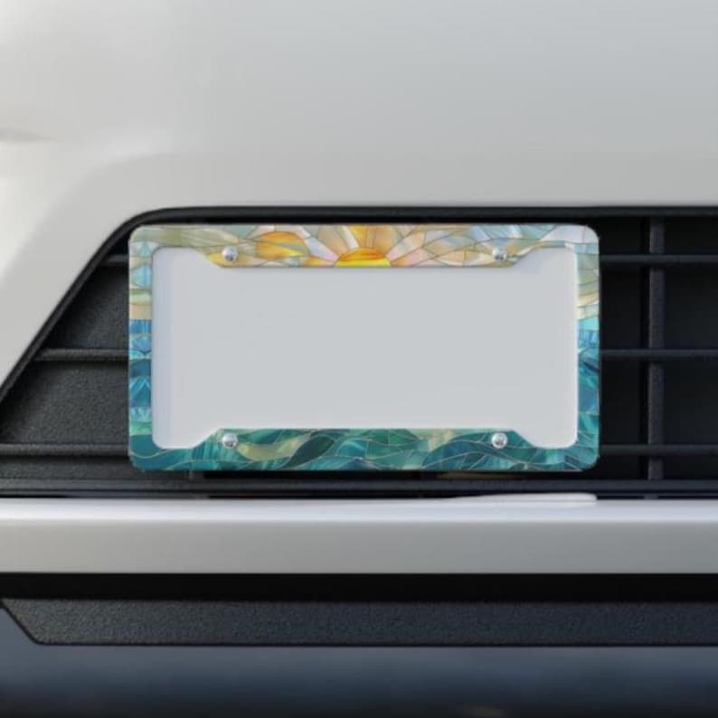 May include: A rectangular license plate frame featuring a stained glass style pattern. The design displays a yellow sun rising over blue and teal waves, with four silver mounting screws visible at the corners of the frame.