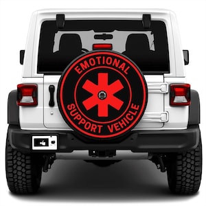 May include: A white Jeep SUV with a black bumper and tires. The spare tire cover is black with red text and a red star-shaped graphic that reads "EMOTIONAL SUPPORT VEHICLE". A license plate with a thumbs-up symbol is visible.
