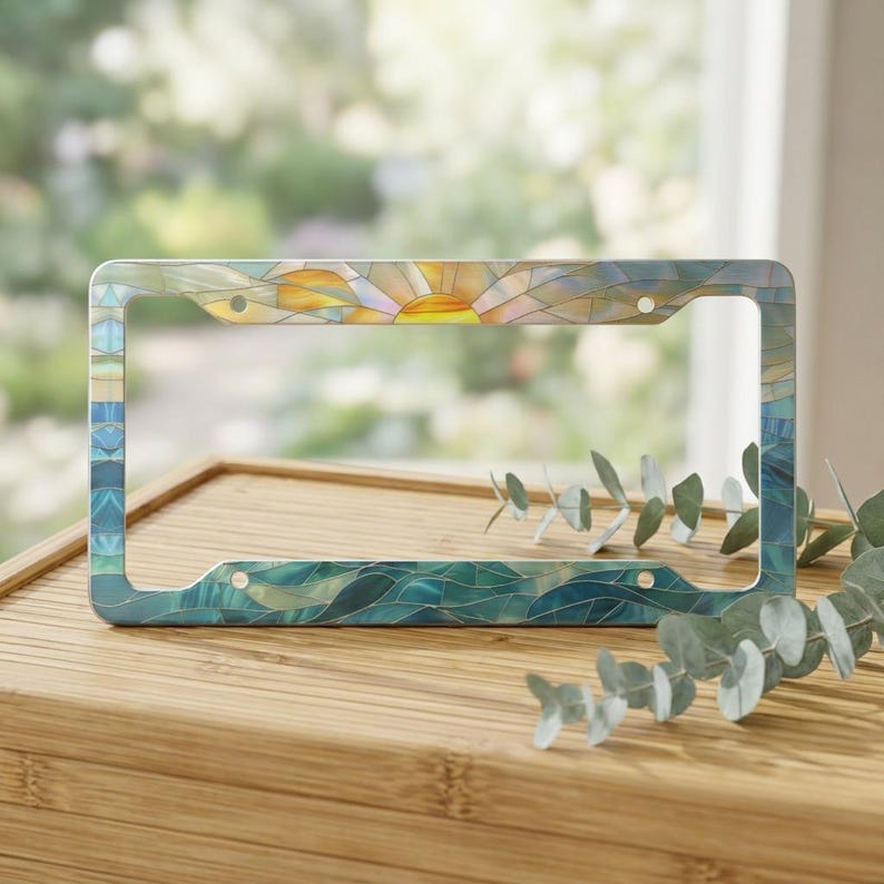 May include: A rectangular license plate frame featuring a stained glass style design. The top section displays a yellow and orange sun rising over a horizon, while the bottom and side sections depict blue and teal waves in a mosaic pattern.