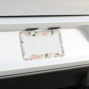 May include: A car license plate frame featuring a repeating pattern of cartoon cats in shades of grey, orange, and white on a light grey background. The frame is rectangular with rounded corners and a clear space for the number plate.