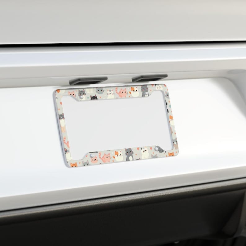May include: A car license plate frame featuring a repeating pattern of cartoon cats in shades of grey, orange, and white on a light grey background. The frame is rectangular with rounded corners and a clear space for the number plate.