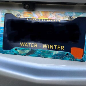 May include: A rectangular license plate frame featuring a stained glass style pattern with a sunrise at the top and blue waves at the bottom. The frame is mounted on a vehicle with a Michigan license plate that displays the text WATER - WINTER.