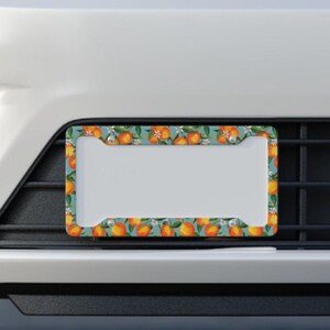 May include: A license plate frame with a teal background and a pattern of oranges, leaves, and white flowers. The frame is rectangular with a white center for the license plate and is mounted on a car.