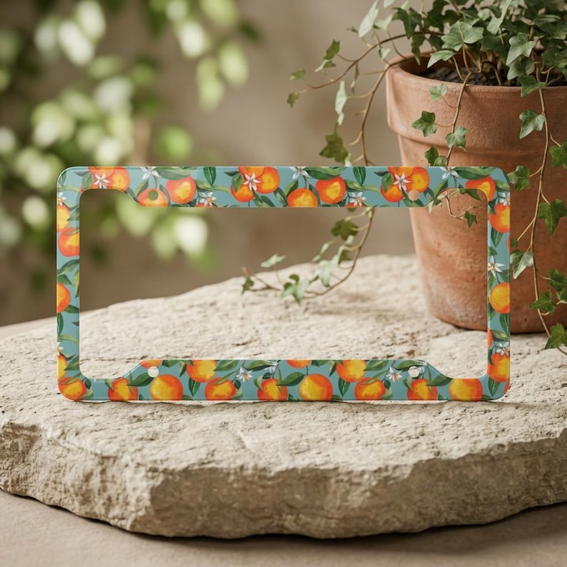 May include: License plate frame with a repeating pattern of oranges, white blossoms, and green leaves on a teal background. The frame is made of metal and has a rectangular shape with rounded corners.