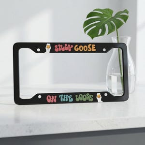 Puede incluir: Marco negro para matrícula con las palabras "SILLY GOOSE" y "ON THE LOOSE" en letras retro coloridas. El marco presenta un gráfico de ganso de dibujos animados. Un artículo decorativo para un vehículo.