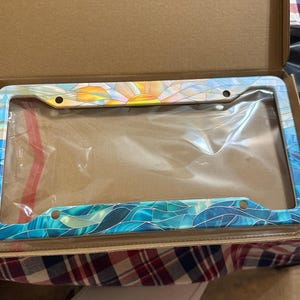 May include: A rectangular license plate frame featuring a colorful stained glass pattern. The top section displays a yellow and orange sunburst design, while the bottom and sides feature various shades of blue and teal wave-like segments.