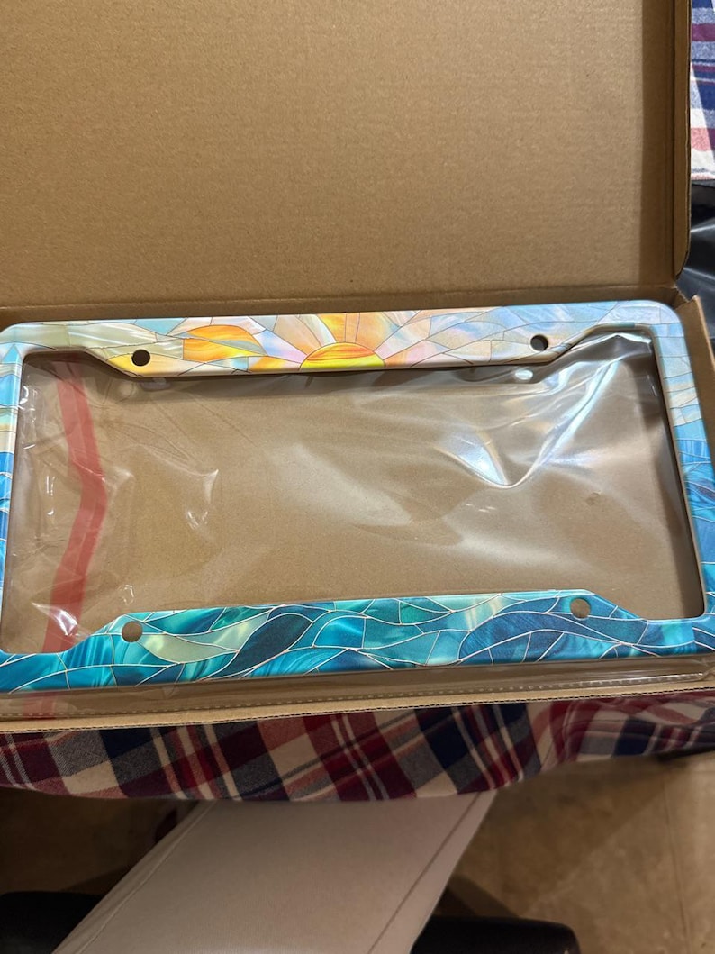 May include: A rectangular license plate frame featuring a colorful stained glass pattern. The top section displays a yellow and orange sunburst design, while the bottom and sides feature various shades of blue and teal wave-like segments.