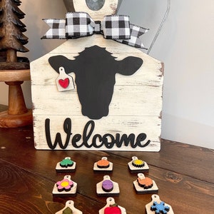 May include: A white wooden sign with a black cow silhouette and the word "Welcome" in black lettering. The sign has a black and white checkered ribbon and a red heart tag. There are 10 small wooden tags with various seasonal designs, including a pumpkin, a flower, a snowflake, and a Christmas tree.
