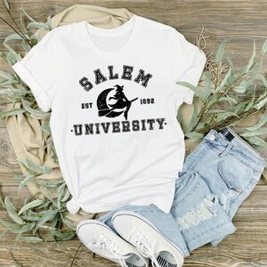 May include: White t-shirt with the text "SALEM UNIVERSITY EST 1692" in black, featuring a witch on a crescent moon. The shirt is paired with light blue denim shorts and white sneakers, arranged on a wooden surface.