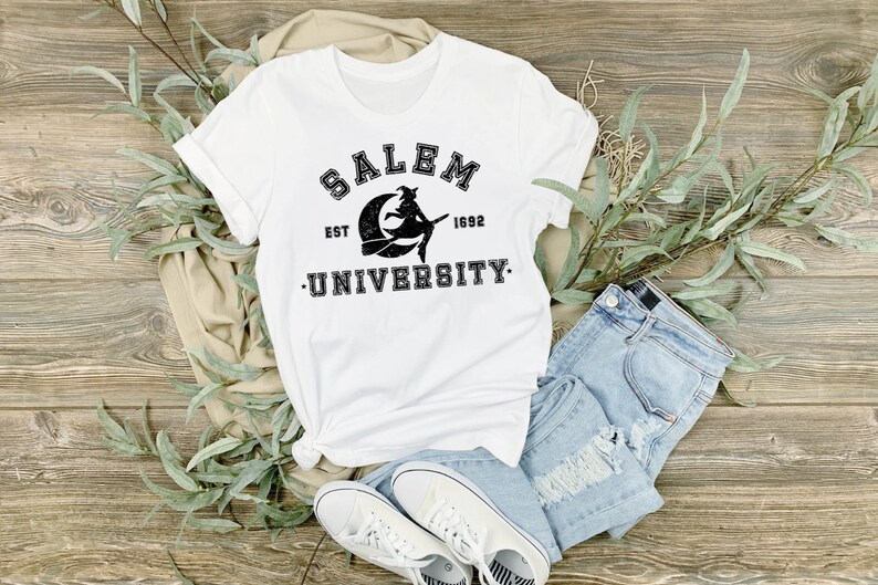 May include: White t-shirt with the text "SALEM UNIVERSITY EST 1692" in black, featuring a witch on a crescent moon. The shirt is paired with light blue denim shorts and white sneakers, arranged on a wooden surface.
