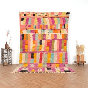 May include: A large, colourful rug with a geometric pattern. The rug features vertical stripes in shades of pink, orange, yellow, green, and blue, with black accents. The rug is displayed against a white wall and wooden floor.