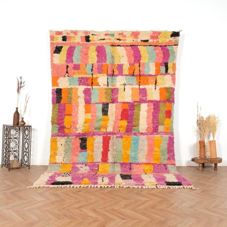 May include: A large, colourful rug with a geometric pattern. The rug features vertical stripes in shades of pink, orange, yellow, green, and blue, with black accents. The rug is displayed against a white wall and wooden floor.