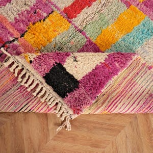May include: A vibrant, geometric rug in shades of pink, yellow, red, and blue. The rug has a textured weave and a fringed edge. The design incorporates a diamond pattern, with a section folded over.
