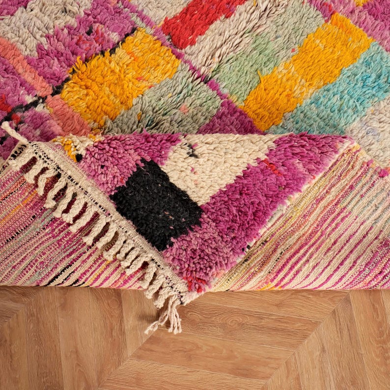 May include: A vibrant, geometric rug in shades of pink, yellow, red, and blue. The rug has a textured weave and a fringed edge. The design incorporates a diamond pattern, with a section folded over.