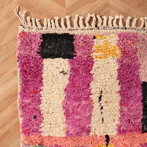 May include: Close-up of a colourful, textured rug with a geometric pattern. The rug features a combination of pink, white, black, and yellow blocks and stripes. The edges are finished with a cream-coloured fringe.