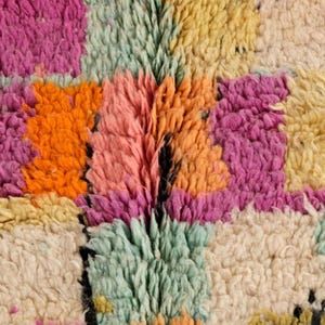 May include: Close-up of a colourful, textured rug. The rug features a patchwork design with squares of orange, pink, yellow, and green. The rug has a shaggy, plush texture, with visible fibres creating a soft, inviting appearance.