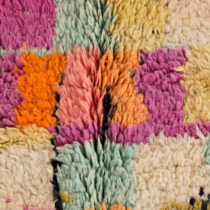 May include: Close-up of a colourful, textured rug. The rug features a patchwork design with squares of orange, pink, yellow, and green. The rug has a shaggy, plush texture, with visible fibres creating a soft, inviting appearance.