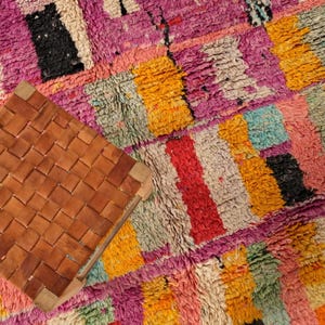 May include: A brown leather stool with a woven seat and a light wood frame is placed on a colourful, textured rug. The rug has a patchwork design with pink, yellow, red, and black, creating a vibrant and artistic look.