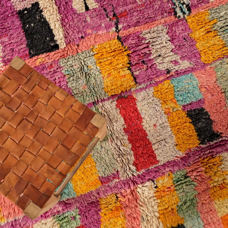 May include: A brown leather stool with a woven seat and a light wood frame is placed on a colourful, textured rug. The rug has a patchwork design with pink, yellow, red, and black, creating a vibrant and artistic look.