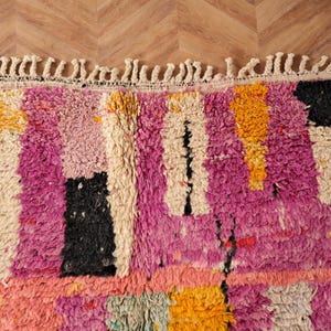May include: Close-up of a colourful, textured rug with a mix of pink, black, yellow, and cream rectangular shapes. The rug has a fringe along the top edge and is made of a thick, plush material. The rug's design is abstract and geometric.