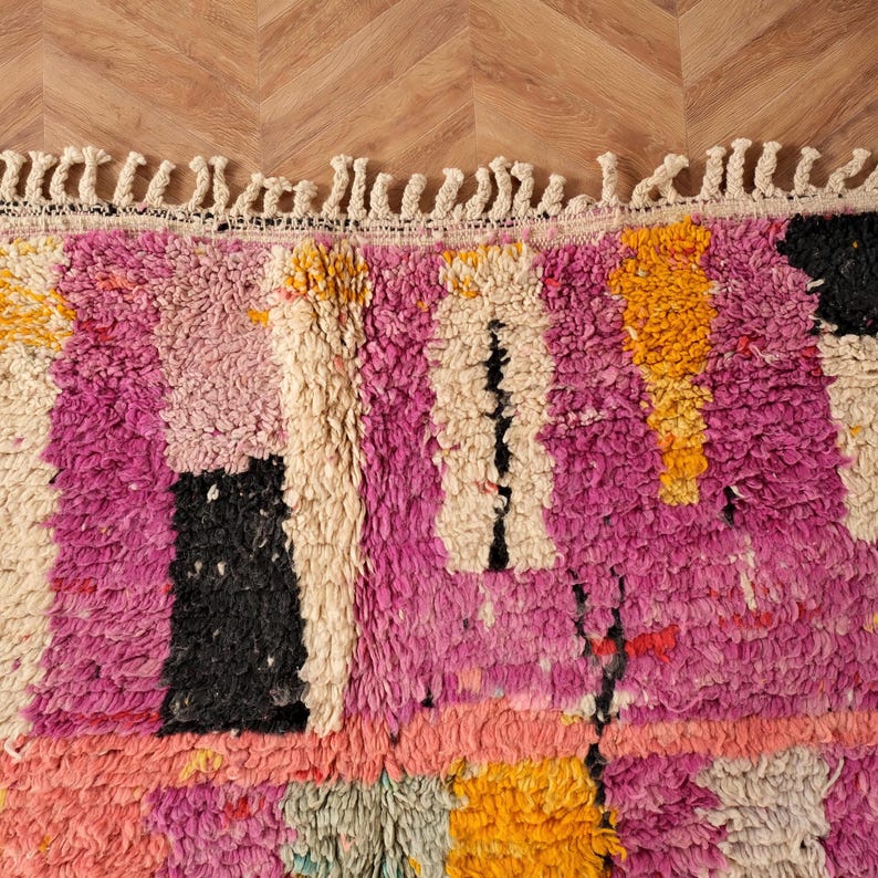 May include: Close-up of a colourful, textured rug with a mix of pink, black, yellow, and cream rectangular shapes. The rug has a fringe along the top edge and is made of a thick, plush material. The rug's design is abstract and geometric.