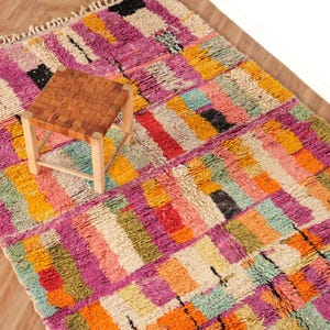 May include: A vibrant, rectangular rug featuring a geometric design in pink, orange, yellow, and green. A small, square stool with a woven leather seat and wooden legs is on the rug. The rug has a fringed border.