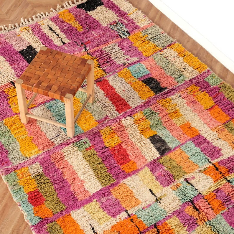 May include: A vibrant, rectangular rug featuring a geometric design in pink, orange, yellow, and green. A small, square stool with a woven leather seat and wooden legs is on the rug. The rug has a fringed border.