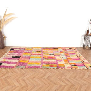 May include: A vibrant, rectangular rug with a geometric design. The rug showcases a patchwork of pink, yellow, orange, and blue blocks. It's made from a thick, woven material and has a fringe along one edge. The rug is displayed on a wooden floor.