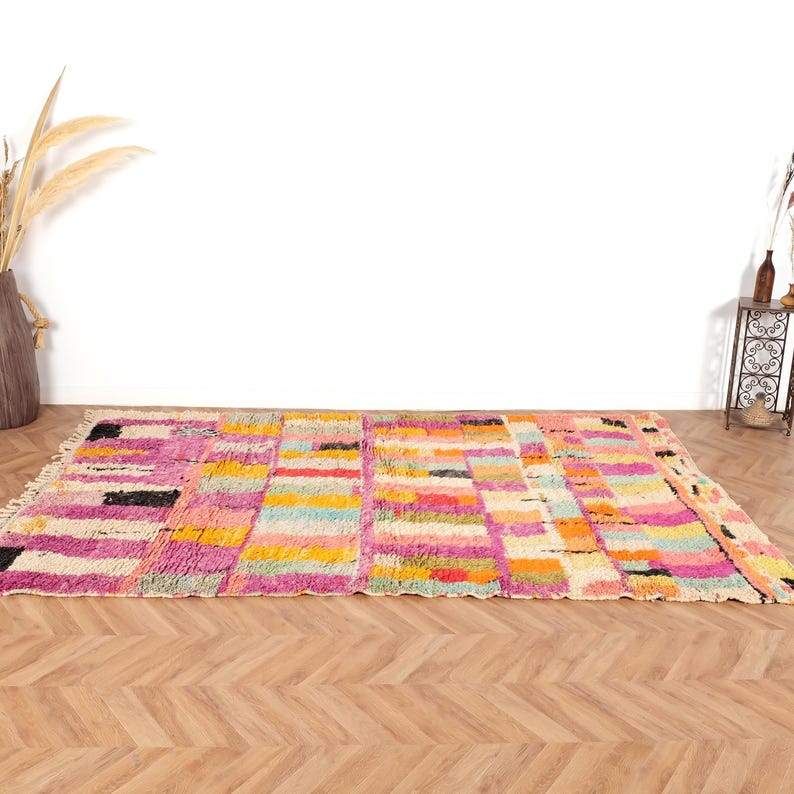 May include: A vibrant, rectangular rug with a geometric design. The rug showcases a patchwork of pink, yellow, orange, and blue blocks. It's made from a thick, woven material and has a fringe along one edge. The rug is displayed on a wooden floor.