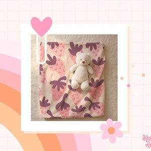 K&ouml;nnte beinhalten: Eine rosa-wei&szlig;e Babydecke mit Erdbeermuster. Ein wei&szlig;er Teddyb&auml;r sitzt auf der Decke.
