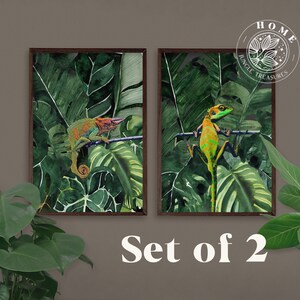 Puede incluir: Conjunto de dos impresiones enmarcadas que presentan vibrantes hojas tropicales verdes con un camaleón y un lagarto posados en ramas. Las impresiones tienen un marco de madera oscura y el texto "HOME JUNGLE TREASURES" está en la esquina superior derecha.
