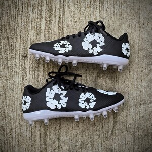 May include: Pair of black cleats with white floral designs. The cleats have white soles and clear studs. Black laces are tied at the top. The cleats are designed for sports.