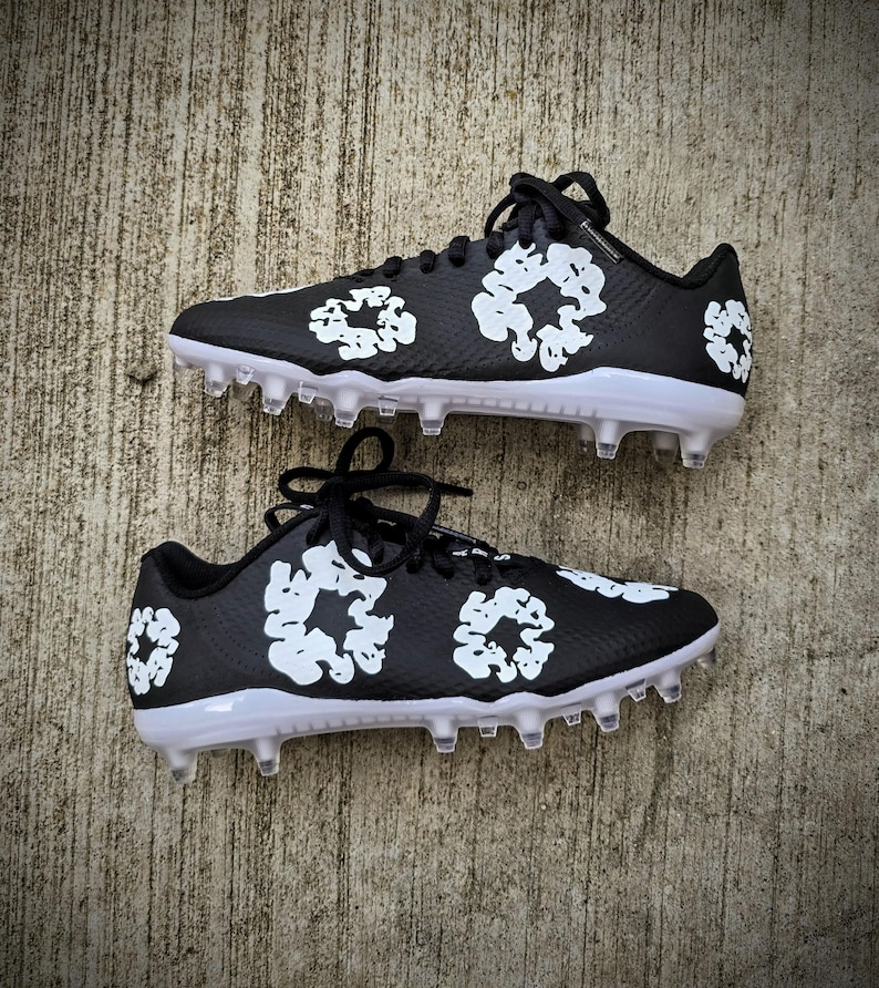 May include: Pair of black cleats with white floral designs. The cleats have white soles and clear studs. Black laces are tied at the top. The cleats are designed for sports.