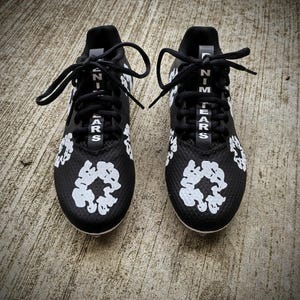 May include: Pair of black athletic cleats with white accents. The cleats feature white floral designs and the text "NIM TEARS" in white. Black laces are tied at the top. The shoes are on a concrete surface.