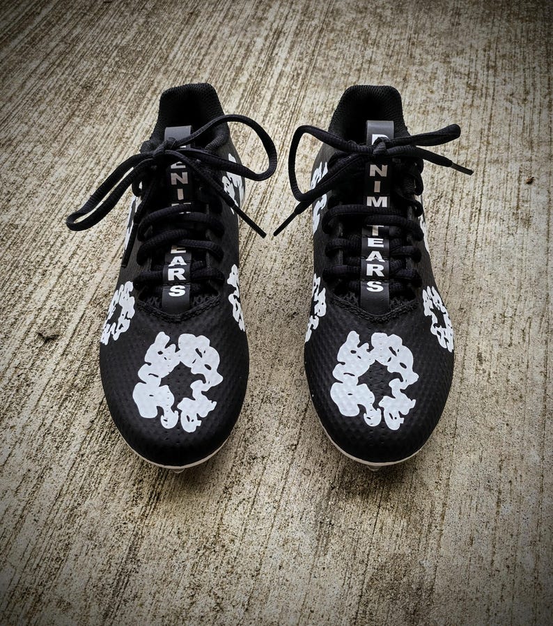 May include: Pair of black athletic cleats with white accents. The cleats feature white floral designs and the text "NIM TEARS" in white. Black laces are tied at the top. The shoes are on a concrete surface.