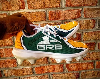 May include: A pair of white, green, and yellow baseball cleats with a logo that says "GRB" and a baseball player graphic.