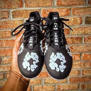 May include: Pair of black athletic cleats with white floral designs and the text "DENIM TEARS" in white. The cleats have black laces and are set against a brick wall background.