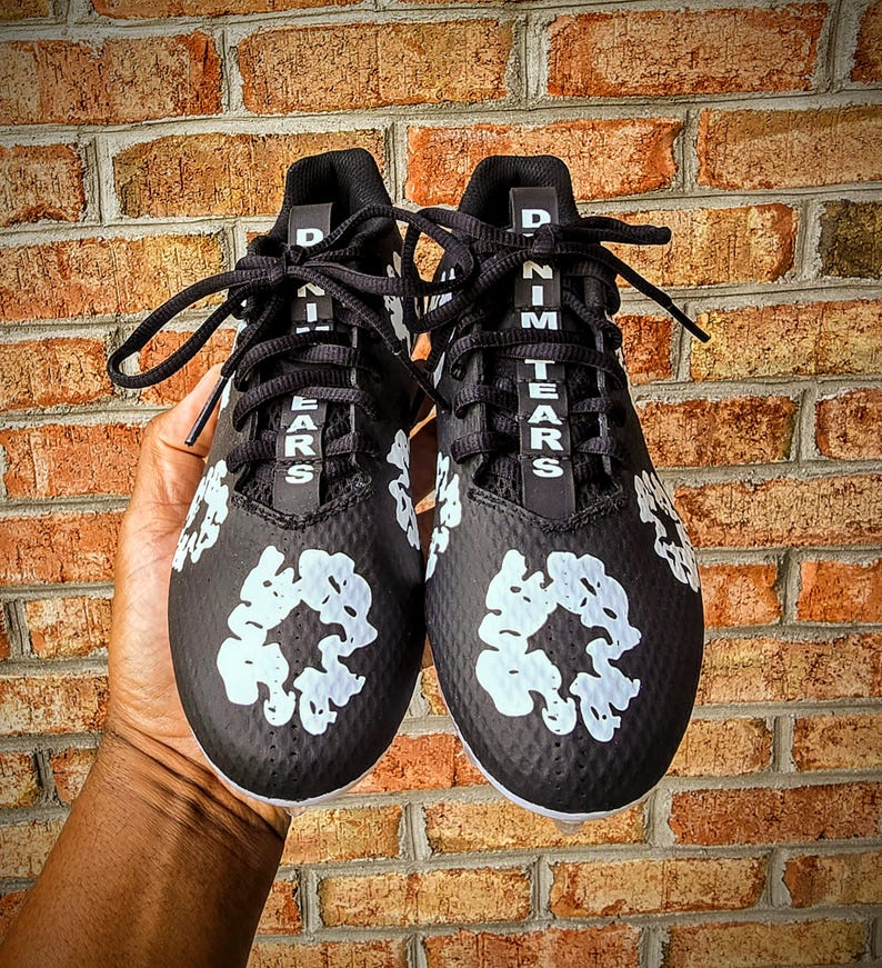 May include: Pair of black athletic cleats with white floral designs and the text "DENIM TEARS" in white. The cleats have black laces and are set against a brick wall background.
