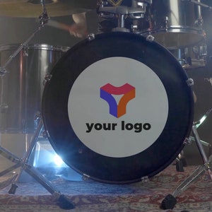 Könnte beinhalten: Eine schwarze Bassdrum mit einem weißen Kreis in der Mitte. Der Kreis enthält ein farbenfrohes abstraktes Logo und den Text "your logo". Die Trommel ist Teil eines Schlagzeugsets mit silbernen Beckenständern und einer silbernen Snare Drum.