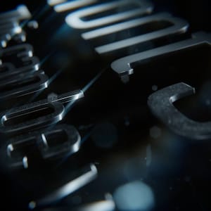 Puede incluir: Primer plano de una superficie oscura con letras metálicas tridimensionales. Las letras son plateadas y parecen ligeramente texturizadas, con un fondo borroso y reflejos. La palabra "JUDGE" es visible.