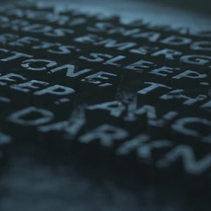 Könnte beinhalten: Nahaufnahme einer dunklen, strukturierten Oberfläche mit eingegrabenen Wörtern. Die Wörter sind "Hords Emerge, Shadows Lorets Sleep. Stone They Creep, Ancient The Darkness."