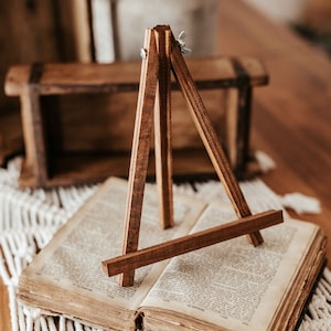 Könnte beinhalten: Ein kleiner Holzstaffelei steht auf einem geöffneten Buch. Die Staffelei ist aus braunem Holz und hat ein schlichtes Design. Das Buch ist alt und hat einen abgenutzten Einband.